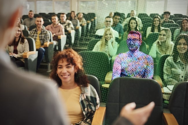 A digitized man wearing glasses sits in a lecture hall surrounded by students, facing a professor speaking. 